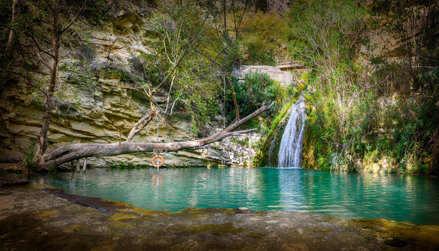 A waterfall dropping down a pale limestone face into a deep turquoise pool, with a fallen tree trunk across the water carrying a rope swing and a lifebuoy, and trees closing over the top