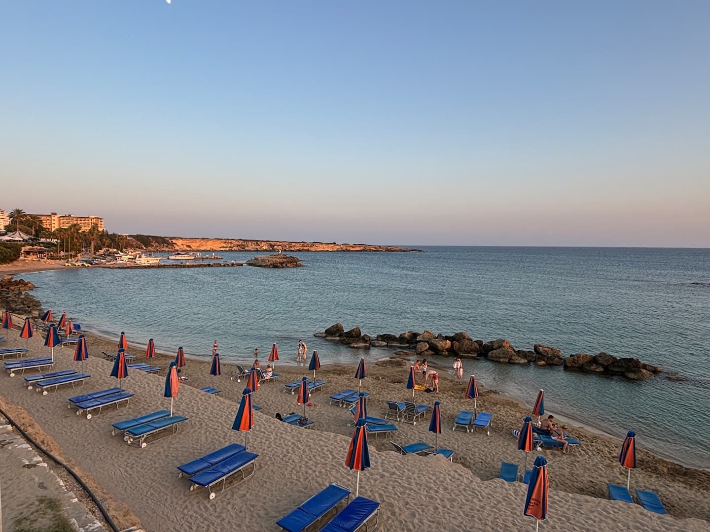 Blue sunbeds and furled parasols on the sand at Corallia, with calm shallow water behind a rock groyne and a low headland beyond